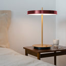 An Umage Asteria Table Lamp with a brass stem and Red shade, placed on a wooden bedside table providing soft illumination.