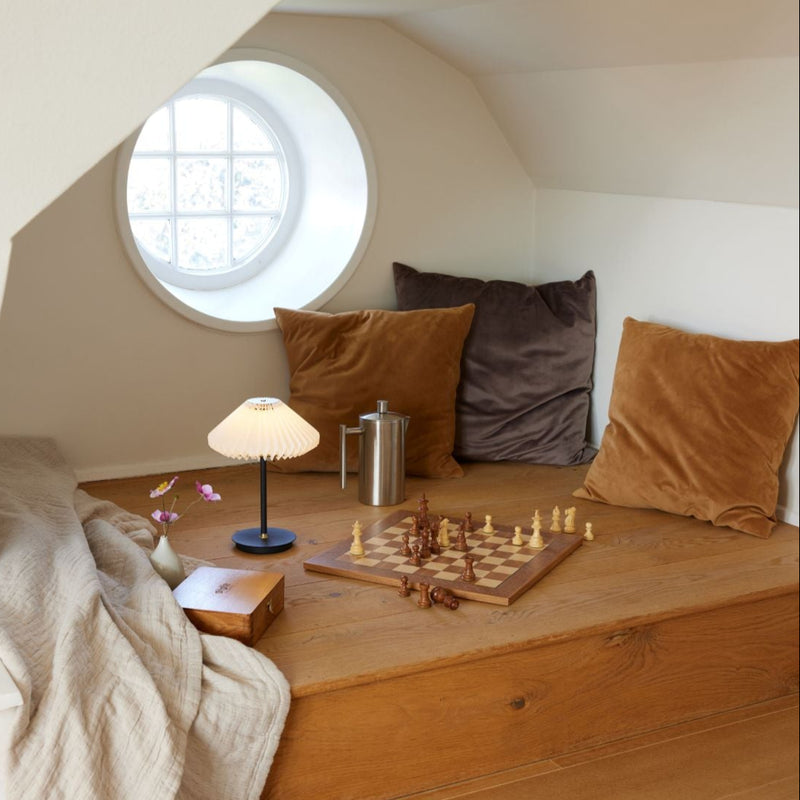 Cozy room with wooden bench, pillows, lamp, and chessboard near a round window.