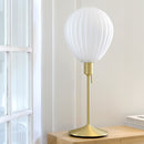 Around the world table lamp by Umage, inspired by the shapes of hot air balloons. The white glass lampshade and elegant Santé table base. A white, globe-shaped table light with a gold-coloured metal stand, placed on a wooden surface next to a window.