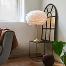 An Eos Feather Table Lamp by Umage with a white lampshade made from goose feathers, mounted on a Santé table base with a USB port, in a room setting.