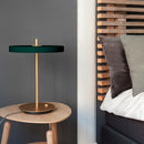 Umage Asteria Table Lamp in Green on a small wooden bedside table next to a grey bed, against a grey wall