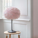 Dusty Pink Umage EOS feather Lampshade on a table lamp agianst a white wood panelled wall on a small side table. The feather lampshade matches the tone of the bare pink wall