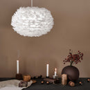 A white feather lampshade by Umage is hanging from the ceiling, surrounded by a cozy setup with candles, a mug, and a decorative vase on a table.