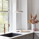 The image shows a modern kitchen setting with two Asteria pendant lights from Umage. The pendants have a minimalistic design with a white diffuser and brass details.